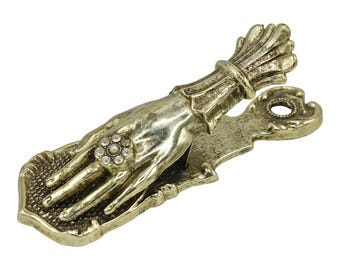 May include: A gold-colored metal clip in the shape of a hand, adorned with a ring of clear stones. The hand holds a bundle of wheat. The clip has an ornate design and is likely used for holding papers or other items.