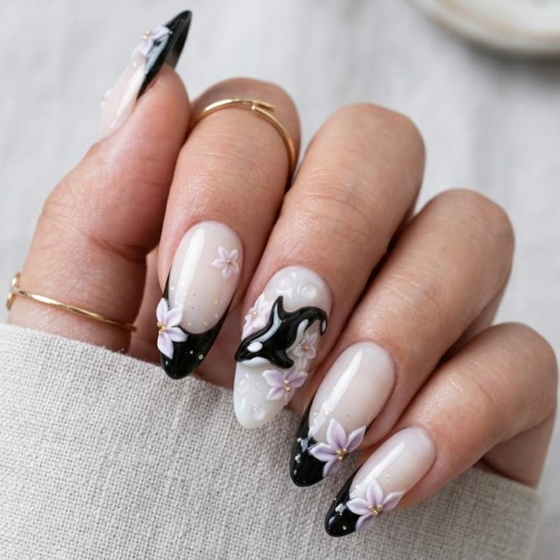 Peut inclure: Ongles de forme ovale avec une manucure fran&ccedil;aise &agrave; pointes noires, orn&eacute;s de fleurs violet clair en relief et d'une petite figurine d'orque noire et blanche sur l'ongle du majeur, le tout sur une base nude transparente.