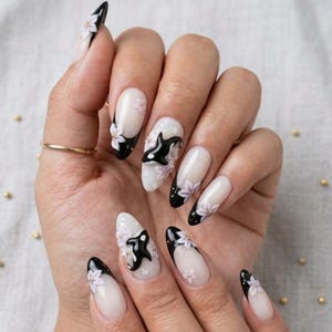 Peut inclure: Ongles en forme d'amande avec une manucure fran&ccedil;aise aux pointes noires sur une base nude transparente. Le design pr&eacute;sente des accents floraux blancs en relief et des silhouettes d'orques noires appliqu&eacute;es sur le vernis &agrave; ongles.