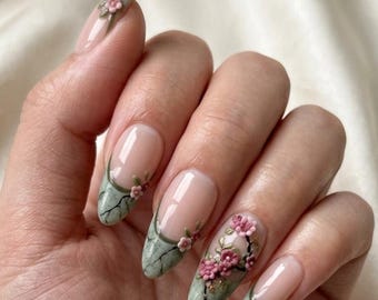 May include: Almond-shaped nails featuring a nude base with sage green French tips. The tips are decorated with black branch-like lines and small, three-dimensional pink floral embellishments that extend onto the nail bed in a delicate pattern.