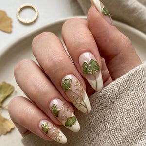 May include: Almond-shaped nails featuring a French manicure base with off-white tips. The nail art includes delicate gold stems and green ginkgo leaf accents painted on the clear and white sections of the nails, creating a botanical design.