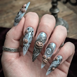 May include: Long, pointed, light blue nail art with metallic silver and copper accents. Each nail features a unique design, including small pumpkins, swirling patterns, and textured elements, creating a Halloween theme.