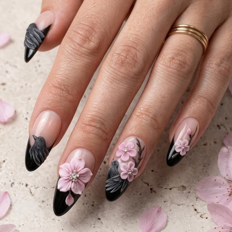 May include: Almond-shaped nails featuring a nude base with black tips. The nail art includes three-dimensional sculpted elements, specifically light pink cherry blossom flowers with small silver centers and dark grey birds perched on branches.