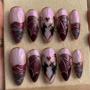 Puede incluir: Un juego de diez u&ntilde;as postizas en forma de almendra con una base rosa claro nacarada. El dise&ntilde;o incluye motivos de alas de murci&eacute;lago en morado oscuro con bordes dorados y siluetas de dos murci&eacute;lagos negros que enmarcan un peque&ntilde;o coraz&oacute;n rojo.
