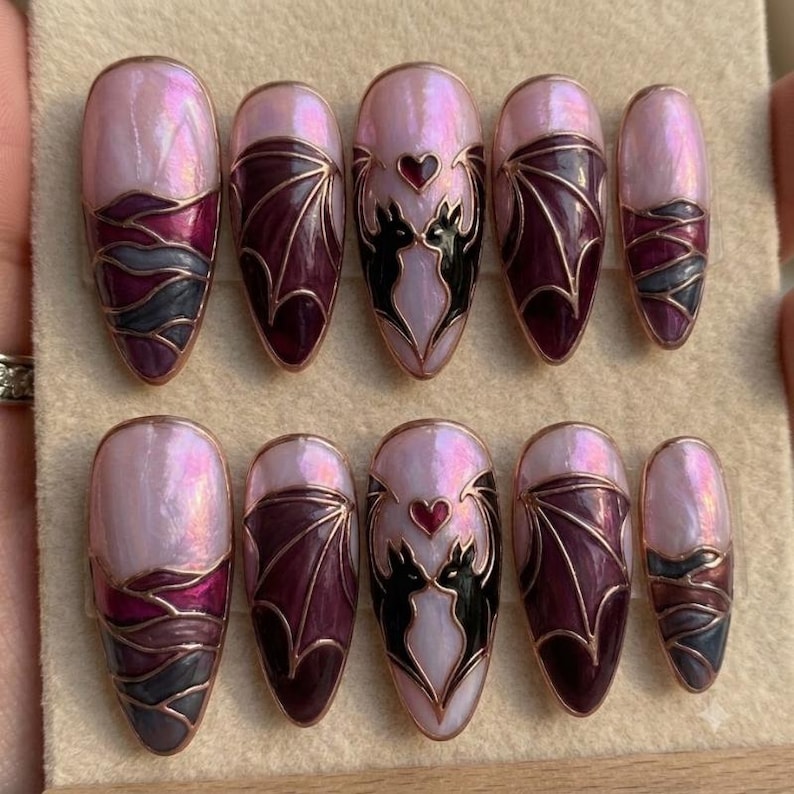 Puede incluir: Un juego de diez u&ntilde;as postizas en forma de almendra con una base rosa claro nacarada. El dise&ntilde;o incluye motivos de alas de murci&eacute;lago en morado oscuro con bordes dorados y siluetas de dos murci&eacute;lagos negros que enmarcan un peque&ntilde;o coraz&oacute;n rojo.
