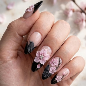 May include: Almond-shaped nails featuring a black French tip design with three-dimensional light pink cherry blossom flowers and small black bird figures. The base of the nails is a natural nude color, accented with tiny silver rhinestones at the center of each flower.