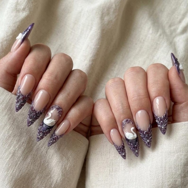 May include: Pointed stiletto nails featuring a nude base with intricate purple lace-like detailing on the tips. Each hand includes an accent nail with a three-dimensional white swan figure surrounded by purple floral patterns and small silver accents.