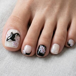 Peut inclure: Ongles d'orteils avec une manucure d&eacute;corative pr&eacute;sentant un motif d'orque noir et blanc sur le gros orteil, agr&eacute;ment&eacute; de fleurs violet clair en relief. Les autres ongles pr&eacute;sentent un m&eacute;lange de noir uni, de blanc transparent et de fleurs.