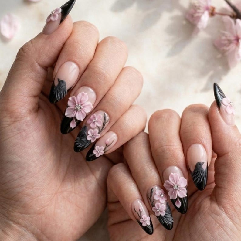 May include: Almond-shaped nails featuring a black French tip design adorned with three-dimensional light pink cherry blossom flowers and small black bird silhouettes. The base of each nail is a natural nude tone, creating a contrast with the dark tips.