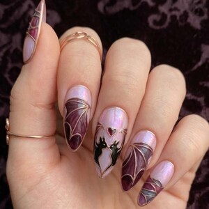 Puede incluir: U&ntilde;as en forma de almendra con efecto de vidriera sobre una base rosa claro iridiscente. Los dise&ntilde;os incluyen alas de murci&eacute;lago de color p&uacute;rpura oscuro delineadas en cobre met&aacute;lico, y una u&ntilde;a central con dos siluetas de murci&eacute;lagos negros flanqueando un peque&ntilde;o coraz&oacute;n rojo.
