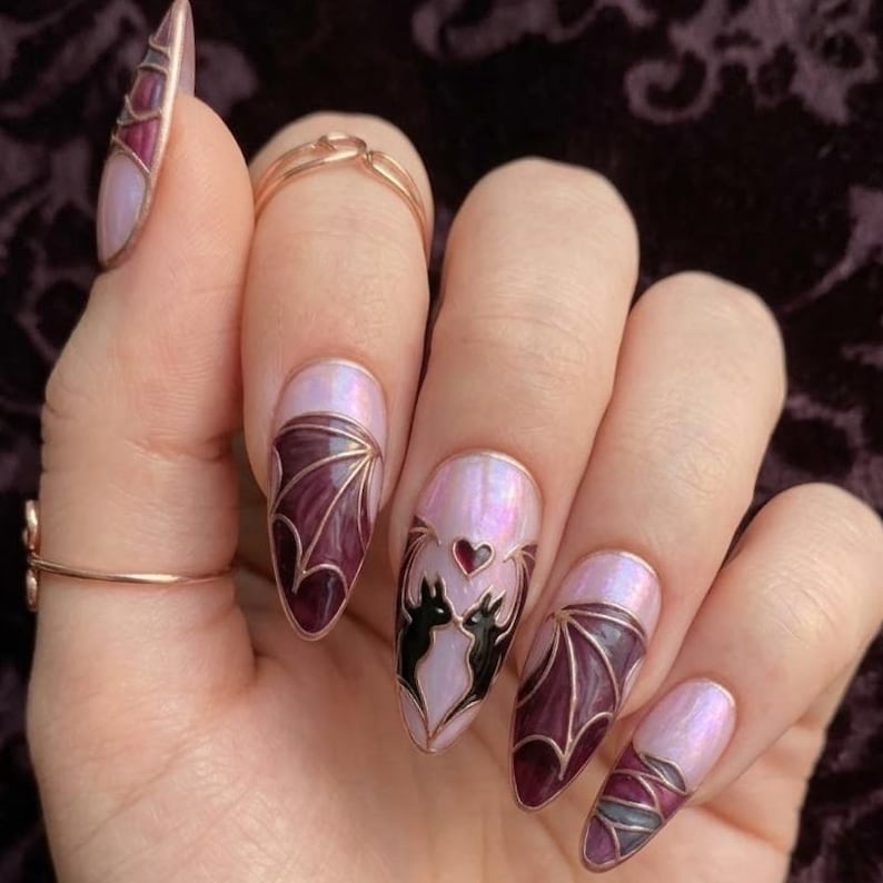 Puede incluir: U&ntilde;as en forma de almendra con efecto de vidriera sobre una base rosa claro iridiscente. Los dise&ntilde;os incluyen alas de murci&eacute;lago de color p&uacute;rpura oscuro delineadas en cobre met&aacute;lico, y una u&ntilde;a central con dos siluetas de murci&eacute;lagos negros flanqueando un peque&ntilde;o coraz&oacute;n rojo.
