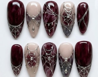 May include: A set of ten almond-shaped press-on nails featuring a mix of deep burgundy marble patterns and pale nude bases. The nails are decorated with intricate silver spider web designs, some accented with small clear rhinestones.