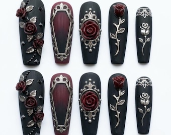May include: A set of ten press-on nails featuring a matte black base with dark red rose embellishments, silver metallic coffin-shaped frames, ornate silver filigree settings, and silver rose stem accents. The designs include studs and floral motifs.