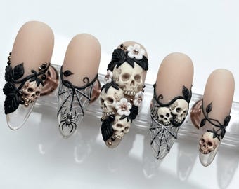 May include: A set of five matte nude-colored artificial nails. Each nail features intricate 3D designs, including skulls, spiderwebs, and floral accents. The designs are in black, white, and gold, creating a gothic and artistic aesthetic.