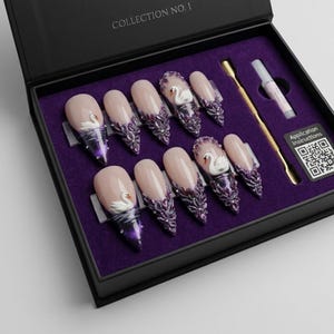 May include: A set of ten press-on nails in a black box labelled COLLECTION NO. 1. The nails feature a nude base with purple tips, intricate floral embellishments, and small white swan figures. Includes a gold cuticle pusher and glue tube.
