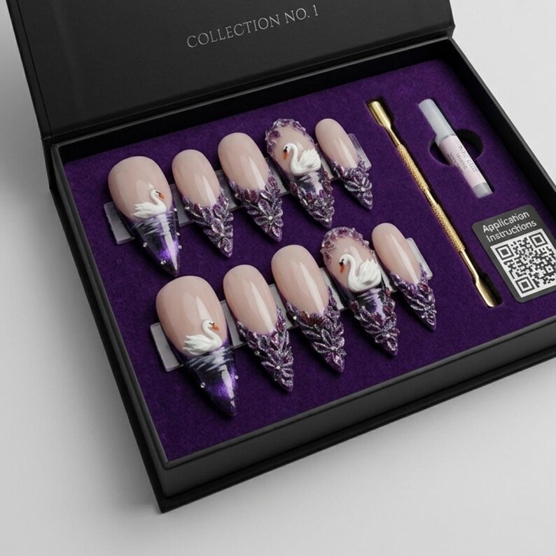 May include: A set of ten press-on nails in a black box labelled COLLECTION NO. 1. The nails feature a nude base with purple tips, intricate floral embellishments, and small white swan figures. Includes a gold cuticle pusher and glue tube.