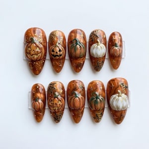 May include: A set of ten almond-shaped press-on nails featuring a glossy amber and brown marbled base. Each nail is decorated with raised 3D pumpkin designs, some carved as jack-o'-lanterns, accented with black spiderwebs, gold stars, and glitter.