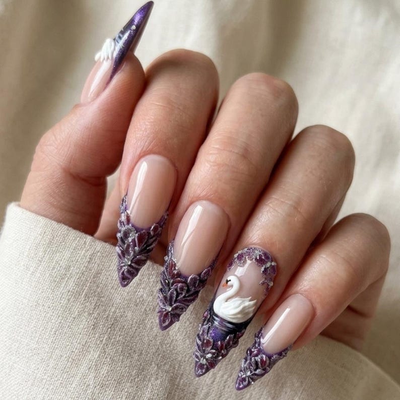 May include: Long stiletto-shaped artificial nails featuring a nude base with deep purple, textured floral embellishments at the tips. One nail displays a small, three-dimensional white swan figure surrounded by purple floral accents and glitter.