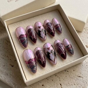 Puede incluir: Un juego de diez u&ntilde;as postizas en forma de almendra con una base rosa claro nacarada. Los dise&ntilde;os incluyen alas de murci&eacute;lago de color morado oscuro, siluetas de dos gatos negros frente a frente y un peque&ntilde;o coraz&oacute;n rojo, delineados con l&iacute;neas doradas.