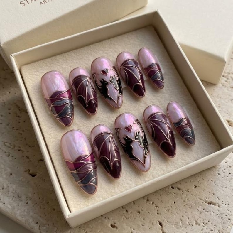 Puede incluir: Un juego de diez u&ntilde;as postizas en forma de almendra con una base rosa claro nacarada. Los dise&ntilde;os incluyen alas de murci&eacute;lago de color morado oscuro, siluetas de dos gatos negros frente a frente y un peque&ntilde;o coraz&oacute;n rojo, delineados con l&iacute;neas doradas.