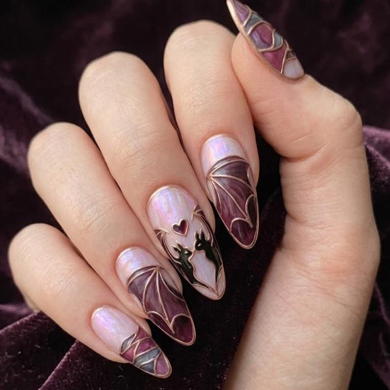 Puede incluir: U&ntilde;as en forma de almendra con base nacarada y contornos met&aacute;licos en relieve. Los dise&ntilde;os incluyen alas de murci&eacute;lago de color p&uacute;rpura oscuro, un peque&ntilde;o coraz&oacute;n y dos siluetas negras de murci&eacute;lagos enfrentados, efecto vitral.
