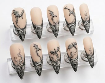 May include: A set of ten stiletto-shaped press-on nails featuring a nude base color. Each nail is decorated with raised, silver-toned relief designs of flying dragons and swirling smoke patterns that extend from the tip toward the center.