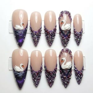 May include: A set of ten almond-shaped press-on nails featuring a nude base with deep purple tips. Some nails are adorned with 3D white swan figures on water, while others feature intricate purple floral embellishments and small silver accents.
