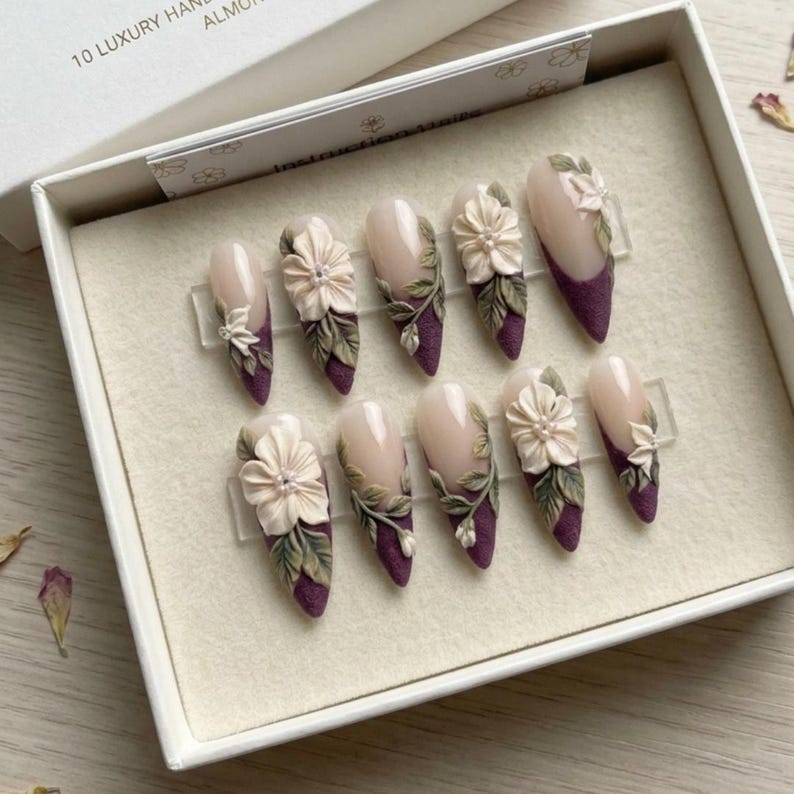 Pu&ograve; includere: Un set di dieci unghie finte a forma di mandorla disposte in una scatola bianca. Ogni unghia presenta una base color carne, una punta color prugna scuro e decorazioni floreali color crema in rilievo con foglie verdi sulla superficie.