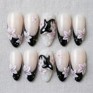 Peut inclure: Un ensemble de dix faux ongles avec une base blanc laiteux et des pointes noires. Le design comprend des fleurs tridimensionnelles violet clair avec des centres dor&eacute;s et deux ongles orn&eacute;s d'une figurine d'orque en relief noir et blanc.