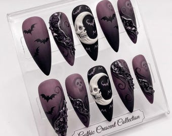 May include: A set of ten long, almond-shaped press-on nails in a clear display case. The nails feature a matte purple-to-black gradient design with gothic embellishments, including bats, skulls, and a crescent moon with a skull. The text "Gothic Crescent Collection" is visible.