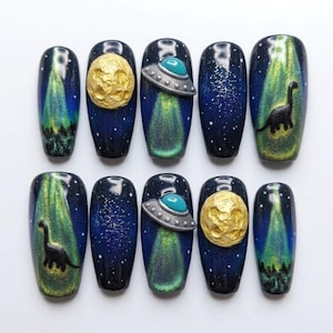 May include: A set of ten press-on nails featuring a space theme with dark blue backgrounds, white star speckles, raised gold textured moons, silver and teal UFOs, and black silhouettes of long-necked dinosaurs standing in green light beams.