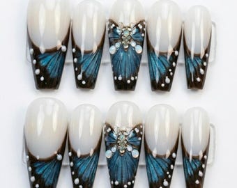 May include: A set of ten press-on nails featuring a long coffin shape with a milky white base and a dark blue butterfly wing pattern at the tips. The design includes small white dots and a central butterfly embellishment with iridescent stones.
