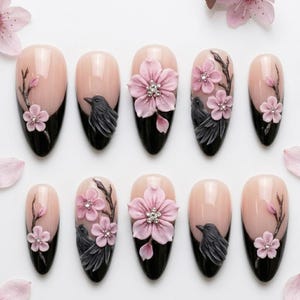 May include: A set of ten almond-shaped press-on nails featuring a nude base and black tips. The designs include three-dimensional light pink cherry blossoms with small clear crystals at the centers, dark grey birds, and brown branches painted on.