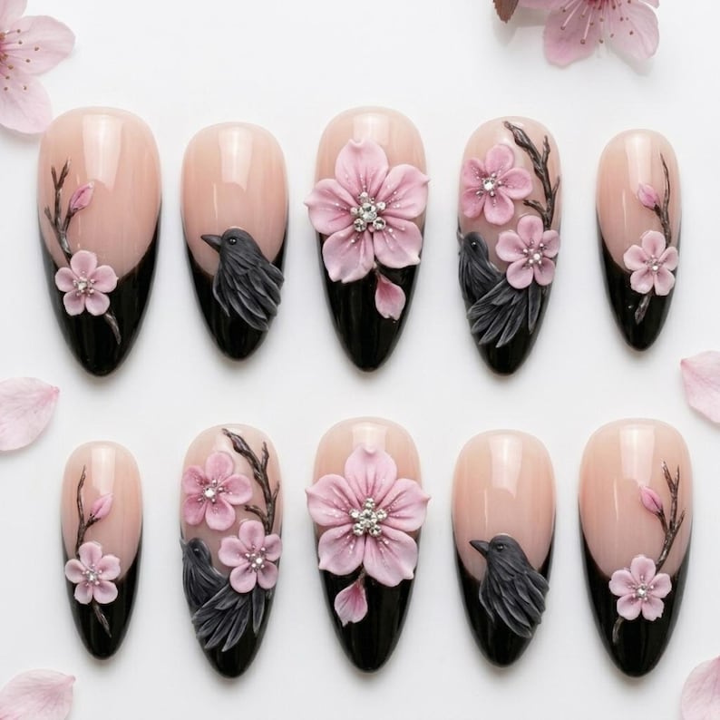 May include: A set of ten almond-shaped press-on nails featuring a nude base and black tips. The designs include three-dimensional light pink cherry blossoms with small clear crystals at the centers, dark grey birds, and brown branches painted on.