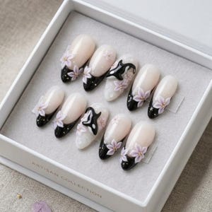 Peut inclure: Un ensemble de dix faux ongles dans une bo&icirc;te blanche marqu&eacute;e Artisan Collection. Les ongles pr&eacute;sentent une base nude brillante avec des pointes noires, orn&eacute;s de fleurs violet clair en relief et de petites figurines d'orques noires et blanches.