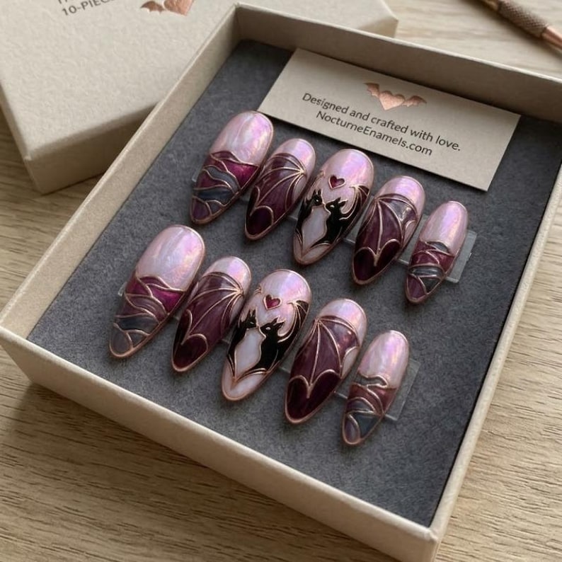 Puede incluir: Un juego de diez u&ntilde;as postizas en una caja de cart&oacute;n, con dise&ntilde;os de esmalte rosa iridiscente y burdeos profundo con bordes dorados. Las u&ntilde;as muestran motivos de alas de murci&eacute;lago y dos u&ntilde;as presentan un par de murci&eacute;lagos negros con un coraz&oacute;n.