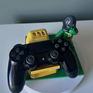 Può includere: Un controller per videogiochi nero poggia su un blocco color oro contrassegnato dal numero 99.0. Accanto al blocco si trova una piccola statuetta verde che indossa una maschera nera e una catena d'oro, su una base quadrata verde e supporto bianco.