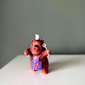 Könnte beinhalten: Eine kleine braune Bärenfigur mit einer weißen Kochmütze und einer violetten Schürze mit weißen Punkten. Der Bär hat eine schwarze Nase und Augen und hält einen kleinen weißen Löffel in der rechten Hand. Die Figur ist glatt modelliert.