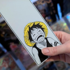 May include: A white rectangular bookmark featuring a smiling cartoon character with a straw hat. The character is depicted in black and white with a yellow hat. The bookmark is held by a hand, with other items in the background.