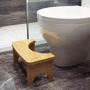 May include: A small, light brown wooden stool with a curved top and four legs. The stool is made of bamboo and is designed to be placed near a toilet. The stool is in a bathroom with a white toilet and grey marble-patterned tile.