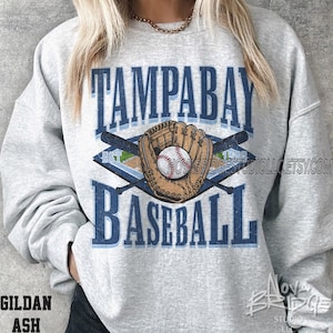 May include: A light gray sweatshirt with a baseball-themed graphic. The design features the text "TAMPA BAY BASEBALL" in blue, along with a baseball glove, baseball, and crossed bats. The sweatshirt also has the text "EST. 1998".