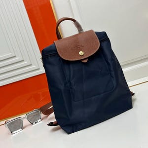May include: A navy blue backpack featuring a brown leather flap with a gold-tone snap button closure and a top handle. The bag has a structured rectangular shape with a smooth fabric finish. A pair of silver-framed sunglasses sits to the left.