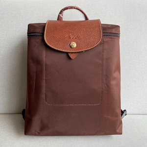 May include: A brown nylon backpack featuring a textured brown leather flap with a gold-tone snap button closure. The bag includes a top handle wrapped in protective plastic, a zippered main compartment, and adjustable shoulder straps on the back.