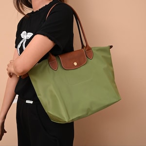 May include: A sage green nylon tote bag featuring brown leather trim, two long shoulder straps, and a fold-over flap closure secured with a gold-tone metal snap button. The bag has a smooth, solid-colored body and a structured leather top flap.