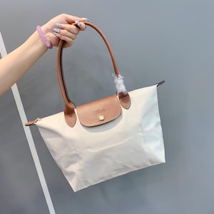 May include: A cream-colored tote bag featuring a brown textured leather flap with a gold-tone snap button closure. The bag includes two long brown leather handles, one of which is partially wrapped in protective plastic, and a top zipper.