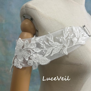 Könnte beinhalten: Weißer Schulterwickel aus floraler Spitze mit zarten Stickereidetails. Befestigt mit einer silberfarbenen Nadel. Der Text "LuceVeil" ist sichtbar. Ein Accessoire für Braut- oder Abendmode.