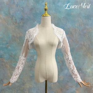 May include: A white lace bolero with long sleeves, displayed on a mannequin. The delicate floral lace design covers the sleeves and shoulders. The text "Luce Veil" is visible in the upper right corner.