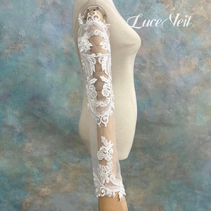 May include: A long, sheer white sleeve accessory featuring intricate white floral lace embroidery. The sleeve is displayed on a mannequin arm and includes the text Luce Veil in the upper right corner, showcasing detailed patterns along the length.