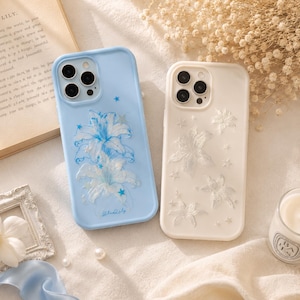 May include: Two phone cases. One is light blue with a floral design and the text "Blue Lily". The other is off-white with a floral pattern. Both cases have camera cutouts and star details.