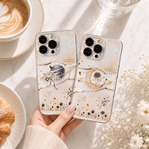 May include: Two clear phone cases featuring celestial illustrations of planets, moons, and roses in soft beige and gold tones. The designs include small stars, floral motifs, and the text Toward the Stars Sound printed in a delicate script.
